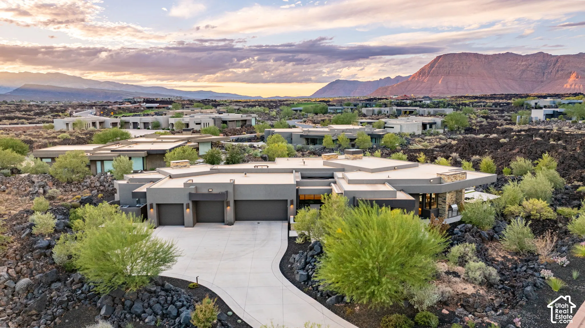 Contemporary residence featuring a multi-car garage, flat roofline, natural stone exterior accents, and a sprawling concrete driveway set against a backdrop of desert mountains