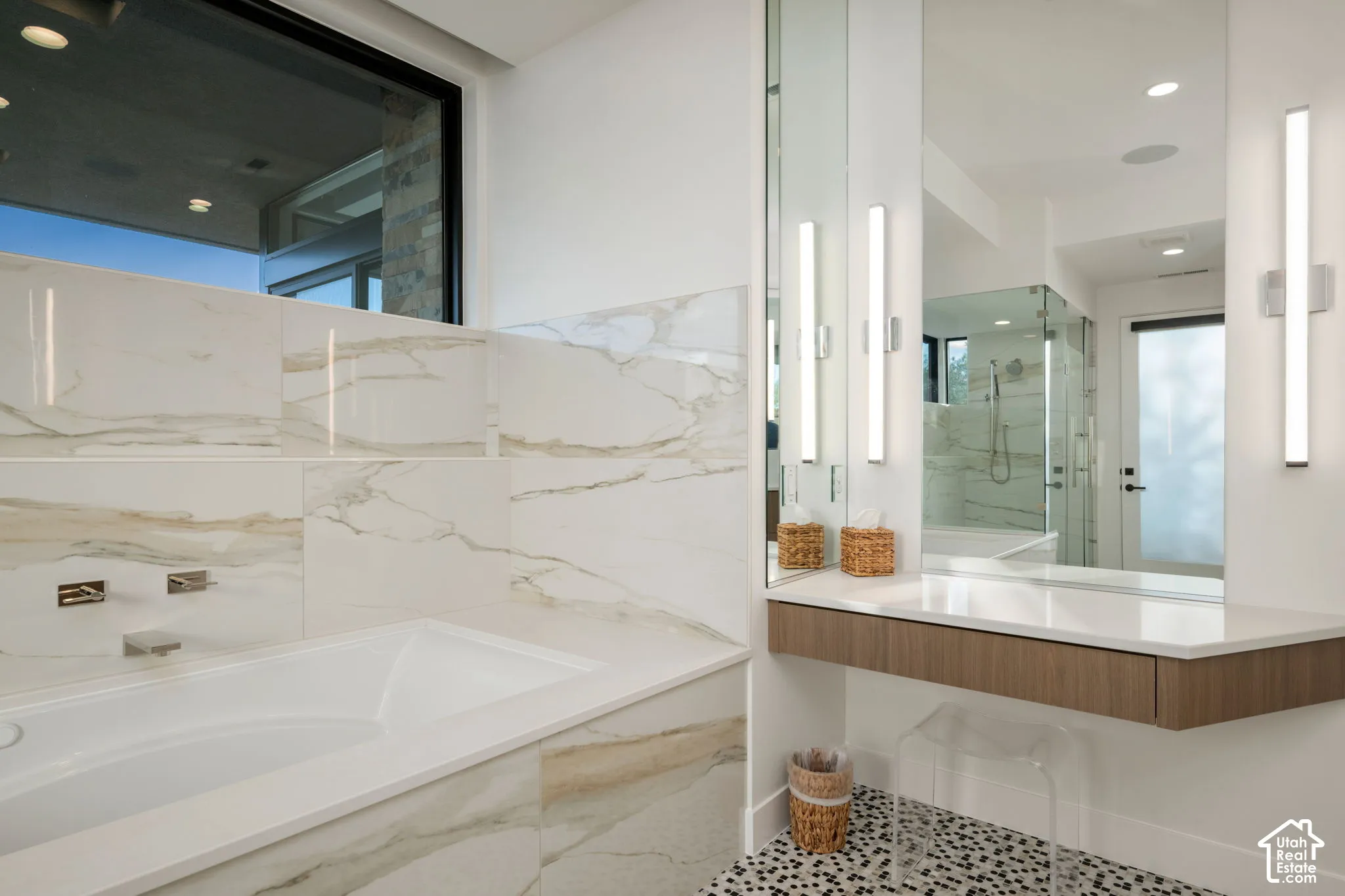 Soaking tub featuring large-format tile surround, wall-mounted polished chrome faucet, floating wood-finish vanity, integrated countertop, and dual linear light fixtures