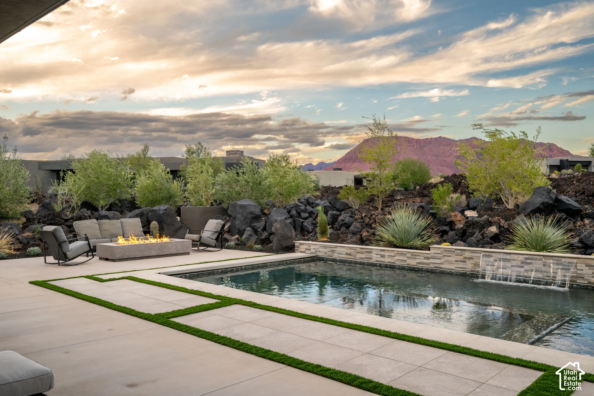 Outdoor living area featuring a rectangular swimming pool, integrated water feature, stone-clad retaining wall, and fire pit set on a concrete patio with artificial turf inlays