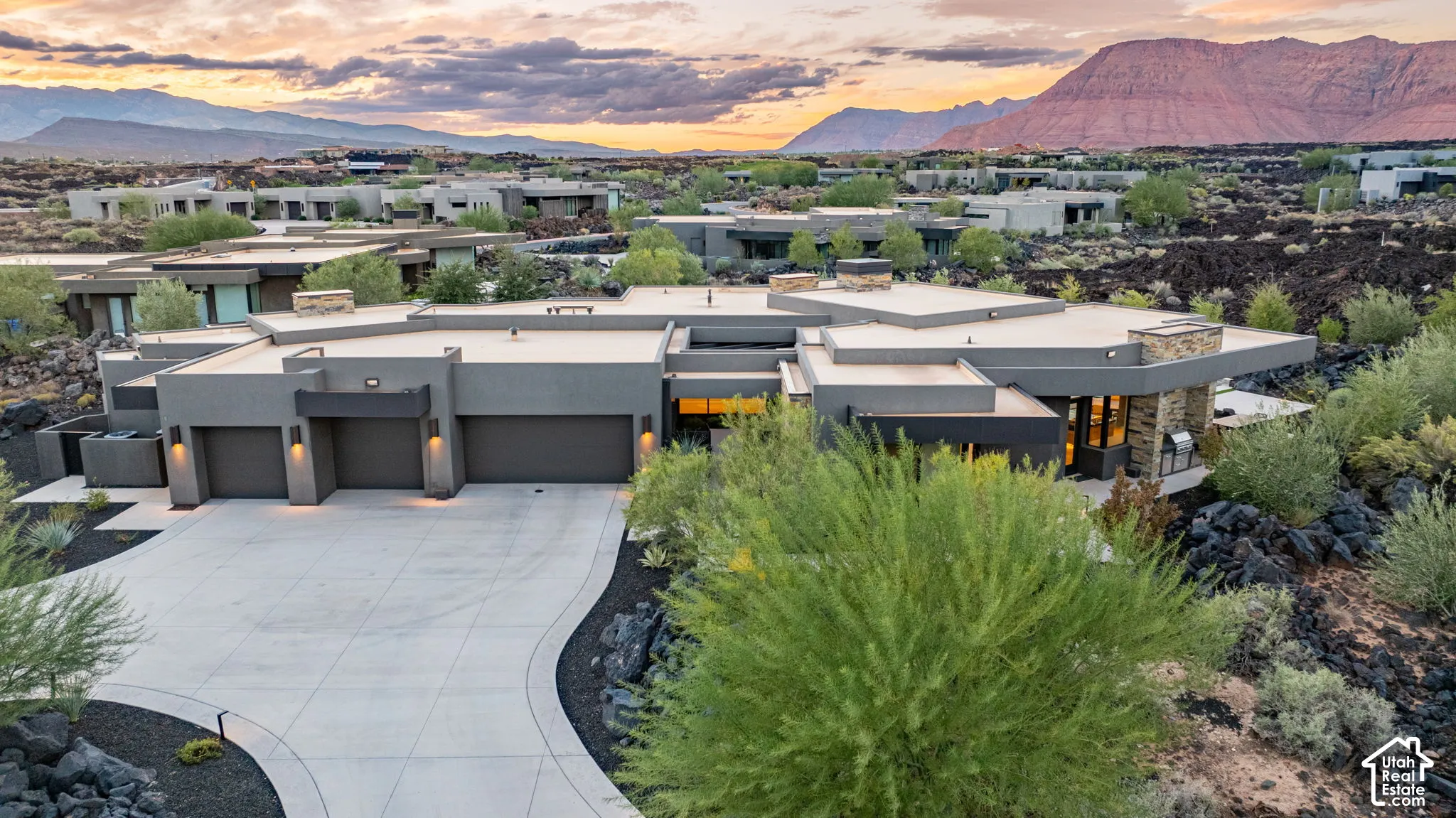 Modern architectural residence featuring a sprawling single-level silhouette, multi-car garage, stone-accented exterior, and expansive paved driveway, set against a backdrop of desert mountains