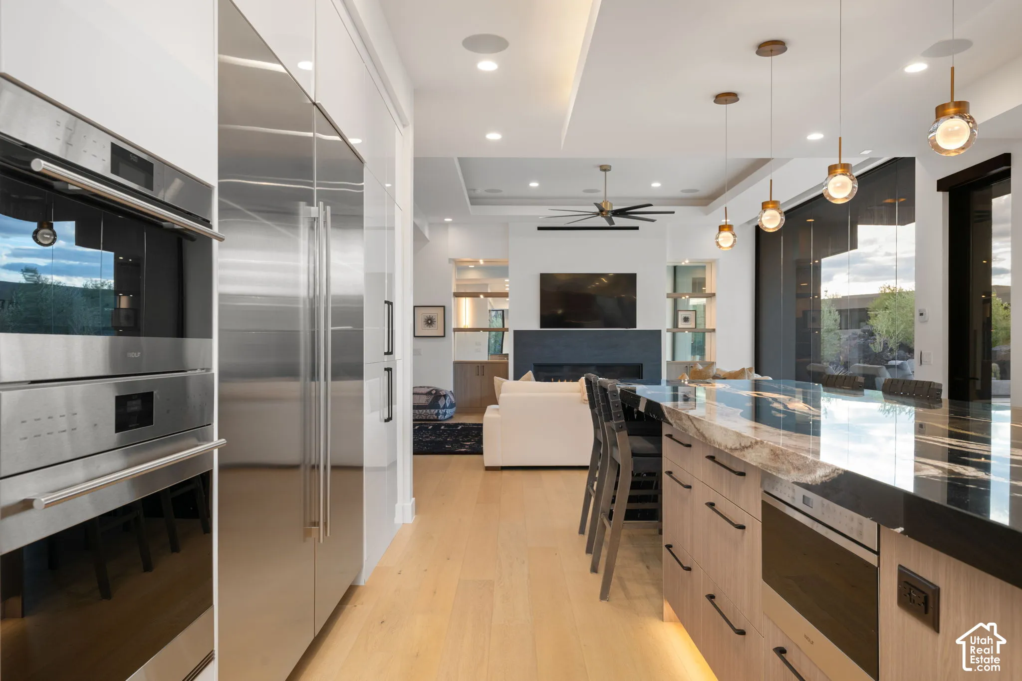 Contemporary kitchen featuring light wood-finish cabinetry, a large stone-topped island with integrated appliances, stainless steel double wall ovens, recessed and pendant lighting, and wood-finish flooring