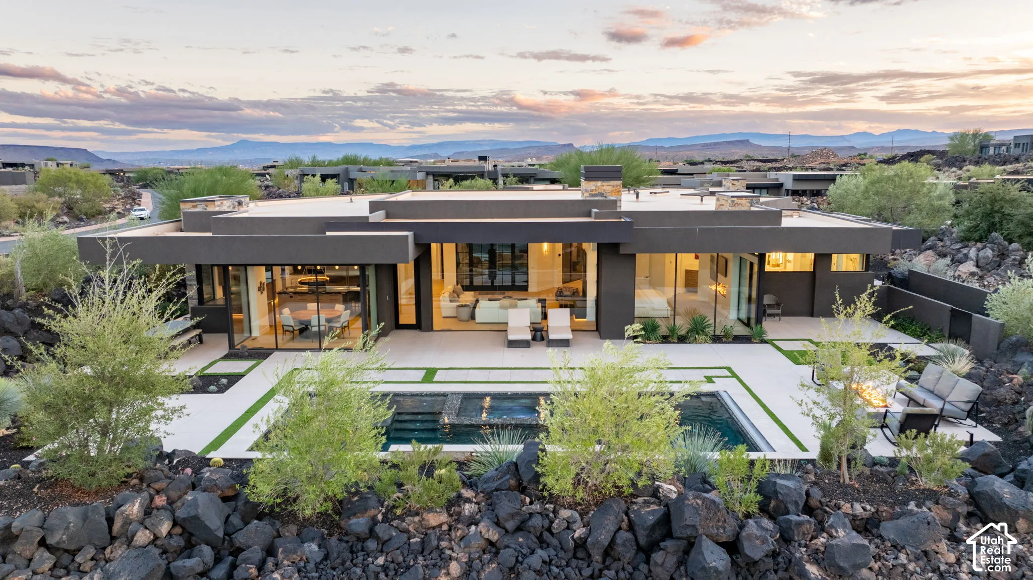 Contemporary single-story residence featuring expansive floor-to-ceiling glass walls, a flat roofline, a built-in outdoor fire pit, and a recessed pool with integrated spa, all set against a desert landscape