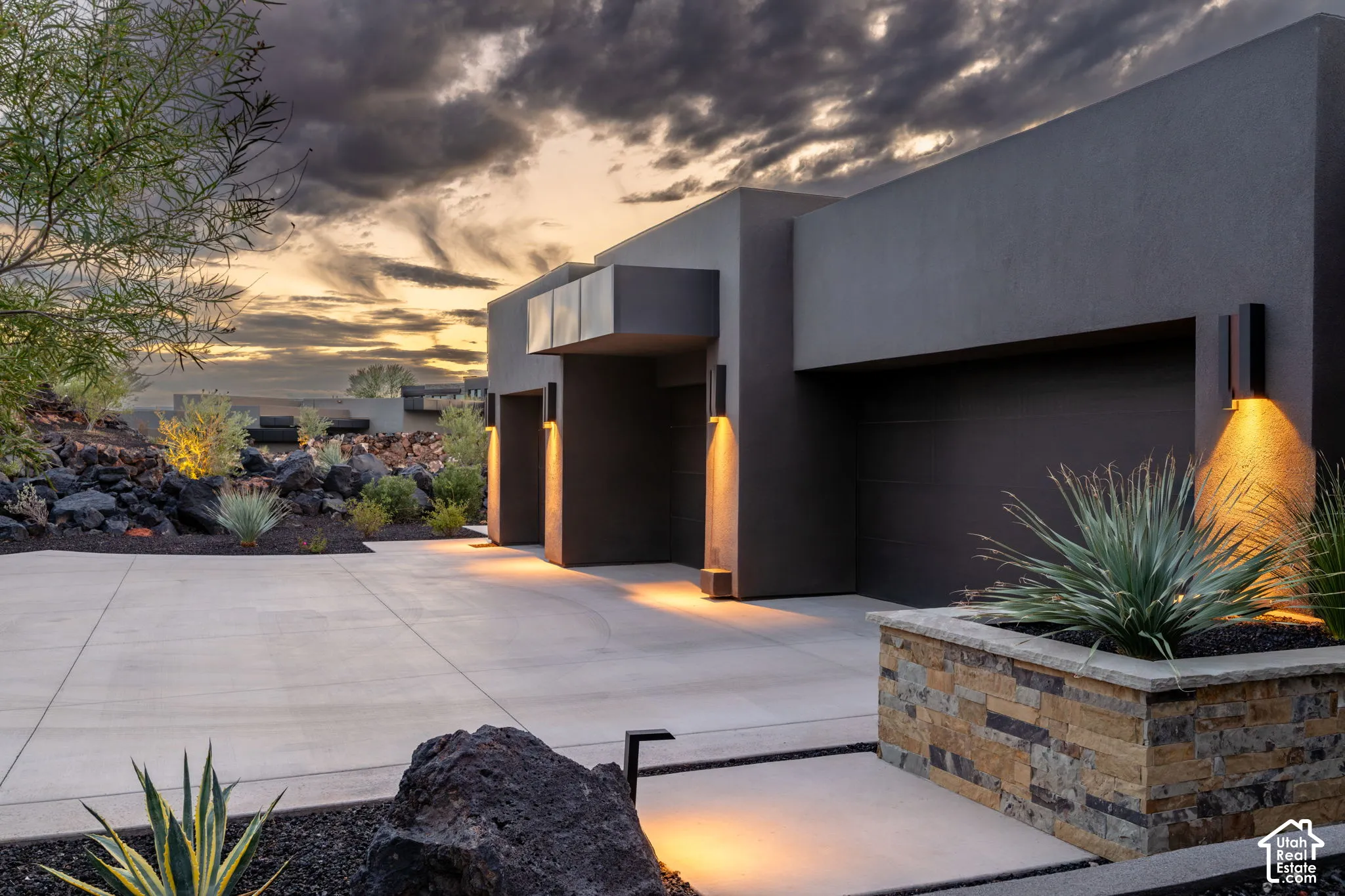 Modern exterior featuring a dark stucco facade, multi-car garage, concrete driveway, stacked stone planter, and integrated wall-mounted lighting