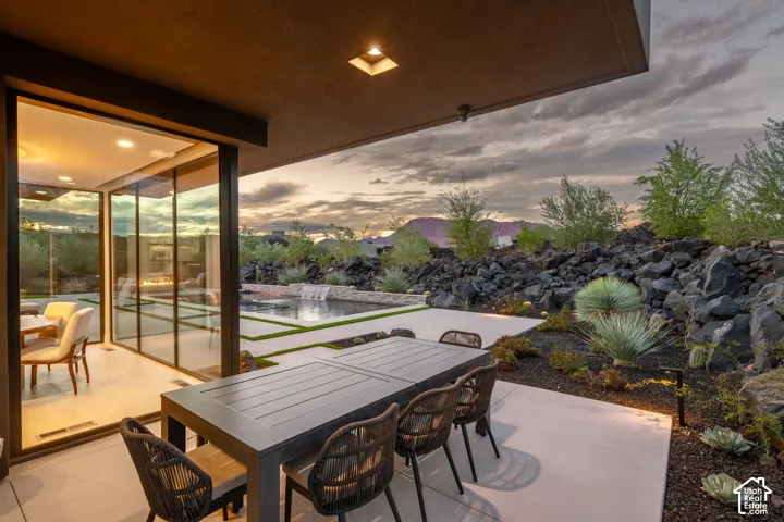 Covered concrete patio featuring floor-to-ceiling glass wall panels, an inground pool with water feature, and desert-inspired landscape with natural rock formations