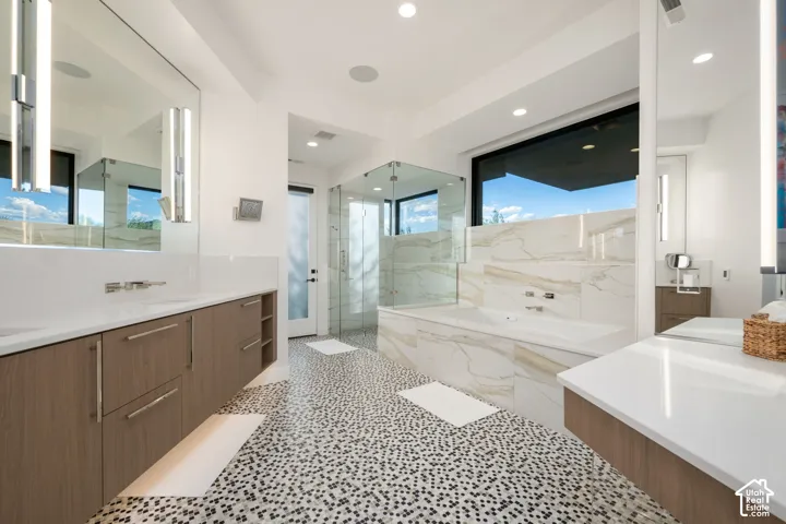 Dual floating vanities with quartz countertops, custom wood-finish cabinetry, floor-to-ceiling stone tilework, glass-enclosed shower, and built-in soaking tub