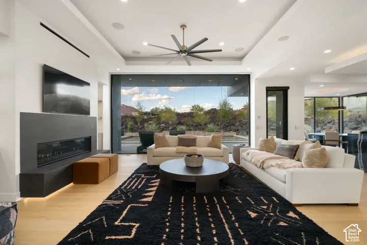 Modern living space featuring wood-finish flooring, a linear fireplace with a stone-finish surround, a tray ceiling with a multi-blade ceiling fan, recessed lighting, and a floor-to-ceiling glass wall