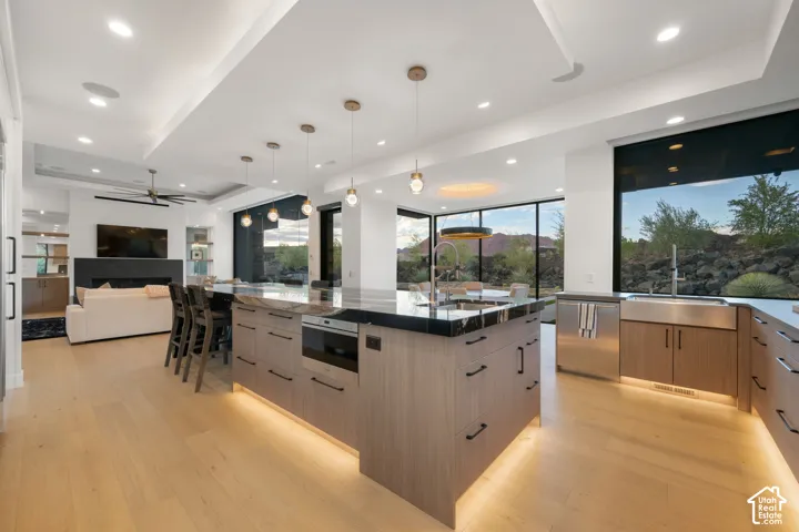 Modern kitchen featuring an oversized island with dark stone countertops, wood-finish cabinetry, recessed ceiling lighting, pendant light fixtures, and stainless steel apron-front sink