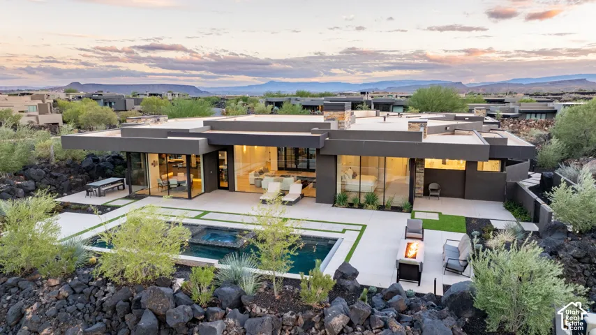 Modern single-story exterior featuring expansive floor-to-ceiling glass walls, integrated concrete patio, negative-edge swimming pool with built-in spa, and stone-accented chimneys