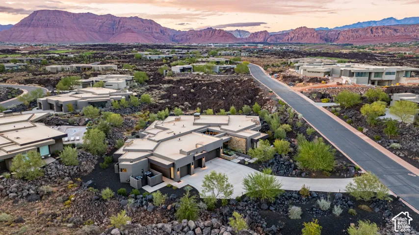Aerial perspective of a contemporary residence featuring a multi-faceted flat roofline, stone veneer accents, integrated multi-car garage, and concrete driveway set against a backdrop of red rock mountain formations