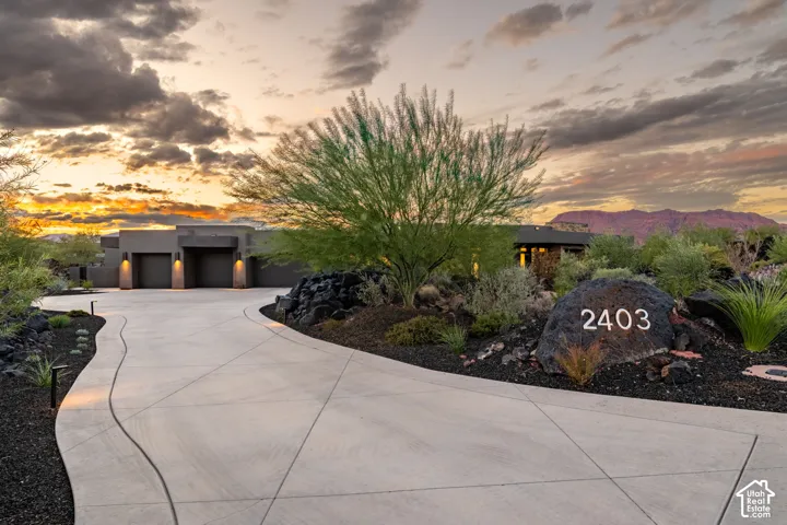 Curved concrete driveway featuring low-maintenance desert landscaping, mature native trees, natural rock formations, and a multi-car garage