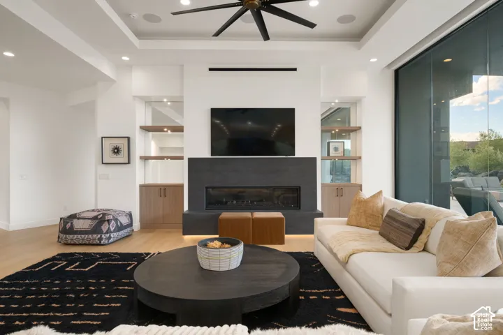 Tray ceiling with recessed lighting and multi-blade ceiling fan, expansive sliding glass wall, floor-to-ceiling fireplace with dark stone surround, and built-in cabinetry featuring floating wood shelving and under-cabinet illumination