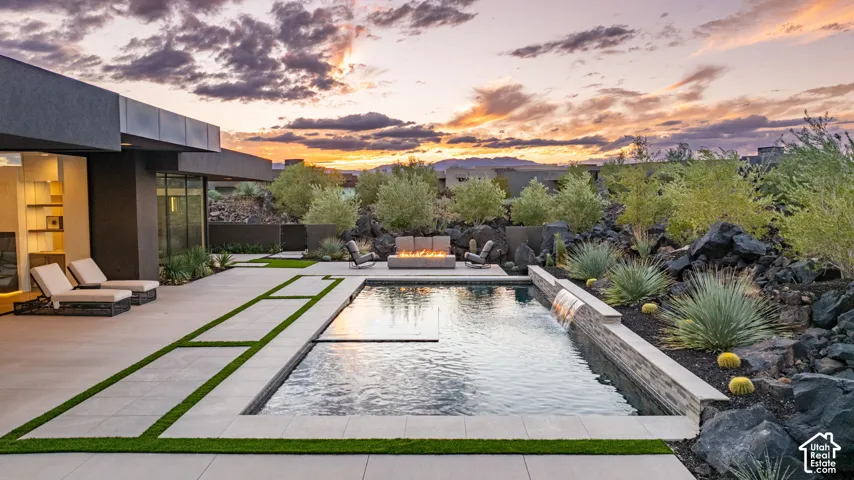 Rear exterior hardscape featuring a rectangular in-ground pool with a stone-clad water feature, integrated turf-accented concrete patio, built-in fire pit, and desert landscaping
