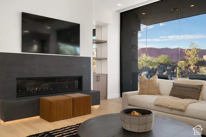 Linear electric fireplace with a dark stone-finish surround, floor-to-ceiling glass wall, built-in cabinetry with illuminated shelving, and light wood-finish flooring