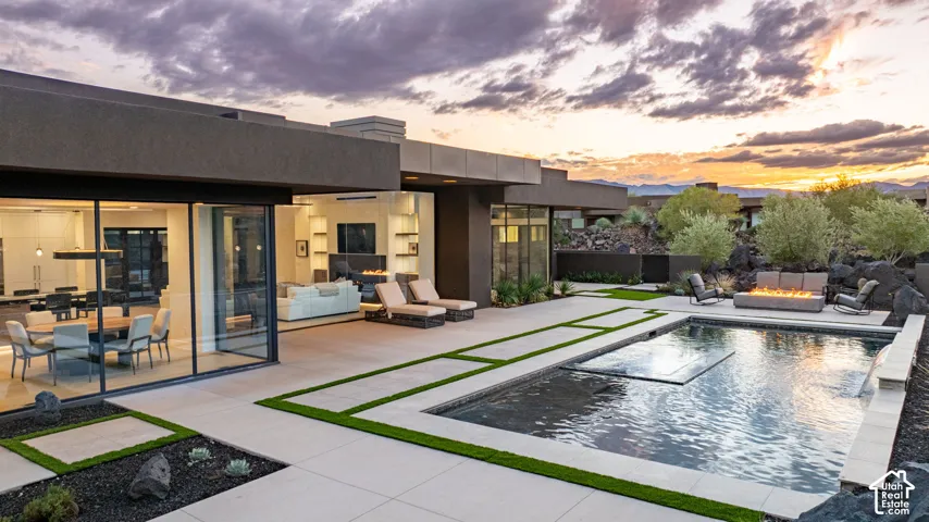 Modern architectural exterior featuring expansive glass walls, a geometric swimming pool with a Baja shelf, structured paver patio with integrated turf, and a built-in outdoor fire pit