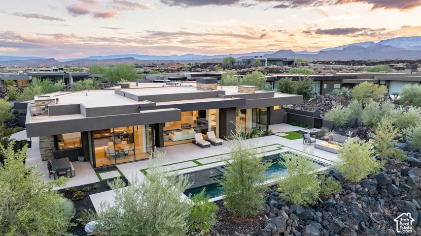 Modern architectural estate featuring an expansive flat roofline, floor-to-ceiling glass walls, integrated in-ground pool, stone exterior accents, and expansive patio space set against a mountainous horizon