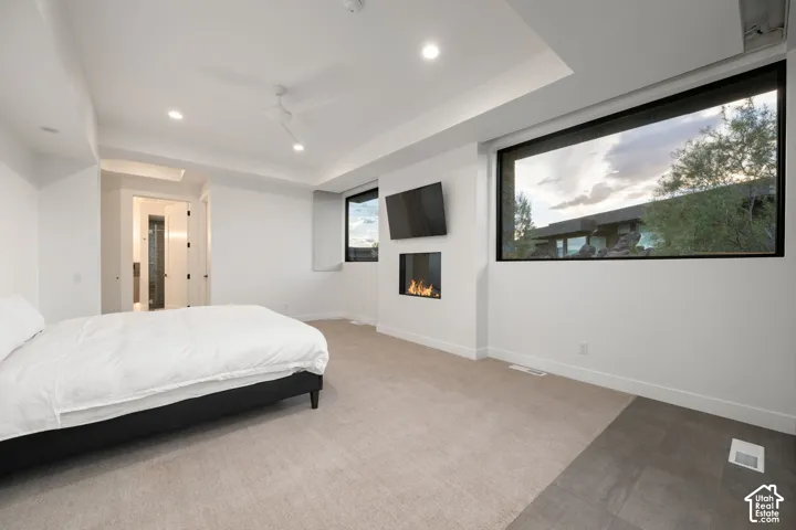 Tray ceiling featuring recessed lighting, wall-mounted linear fireplace, expansive black-framed windows, and neutral carpeting