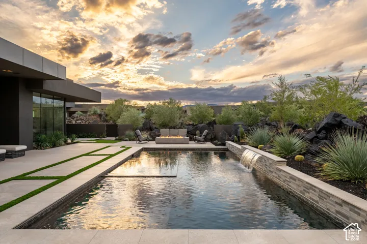 Outdoor living space featuring a custom pool with a waterfall feature, built-in stone-accented fire pit, expansive concrete patio with integrated turf accents, and desert landscaping