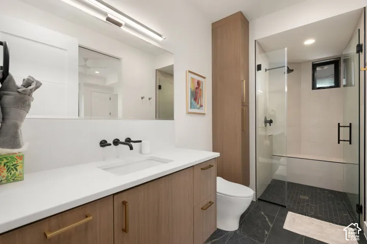 Wood-finish cabinetry with brass hardware, quartz countertop, wall-mounted matte black faucet, full-height storage tower, and walk-in shower with stone tile flooring