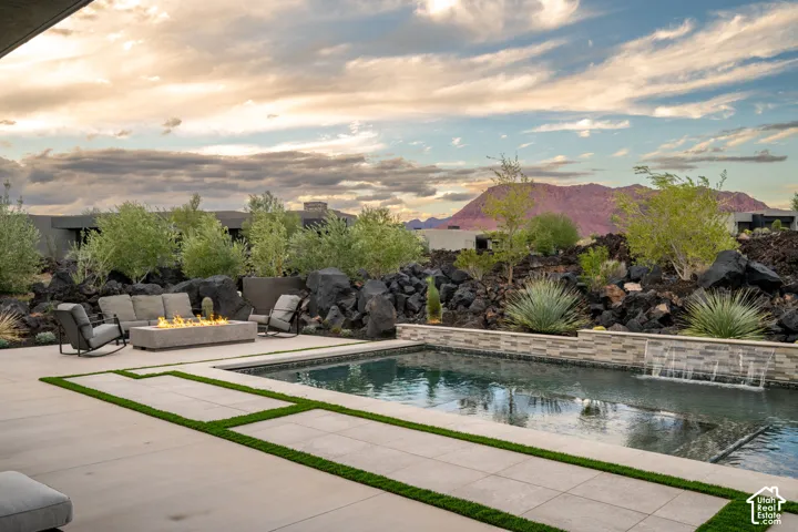 Outdoor living area featuring a rectangular swimming pool, integrated water feature, stone-clad retaining wall, and fire pit set on a concrete patio with artificial turf inlays