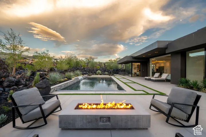 Modern outdoor living area featuring a rectangular concrete fire pit, a linear swimming pool with an integrated waterfall feature, expansive stone paver patio, and natural volcanic rock landscaping