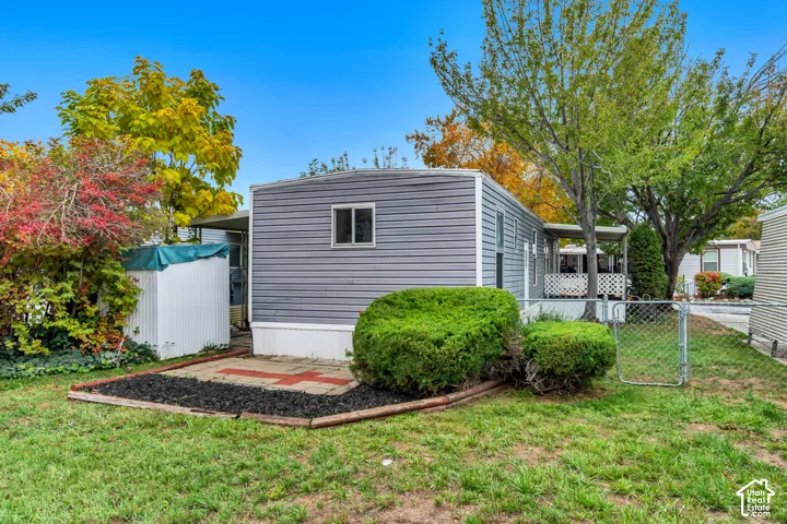 Exterior featuring gray horizontal siding, an attached metal carport, a stone-paved patio area, and chain-link perimeter fencing