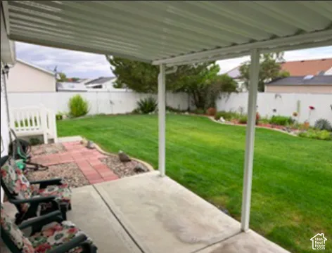 Covered patio featuring a concrete slab surface, metal support columns, enclosed backyard with white vinyl fencing, manicured lawn, and stone-bordered planting beds
