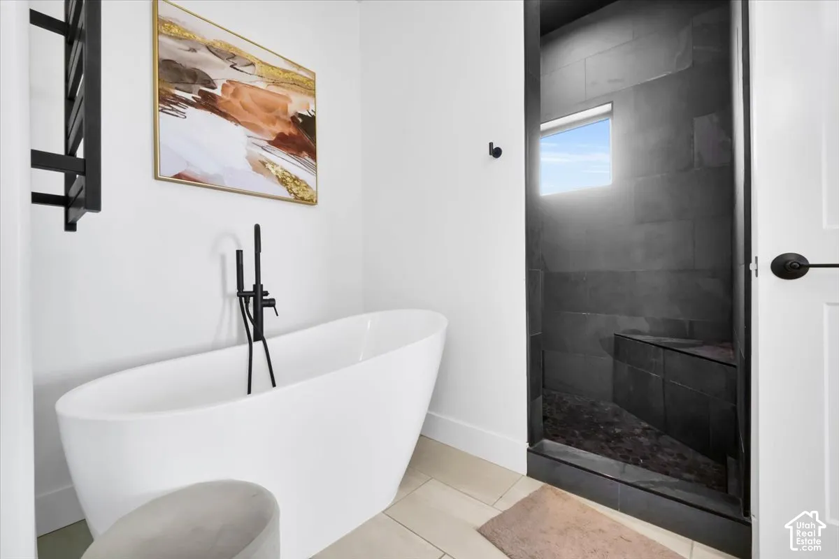 Freestanding white soaking tub with matte black floor-mounted faucet and matching wall-mounted towel warmer, paired with a walk-in shower featuring dark tile surround, built-in bench seating, and square transom window