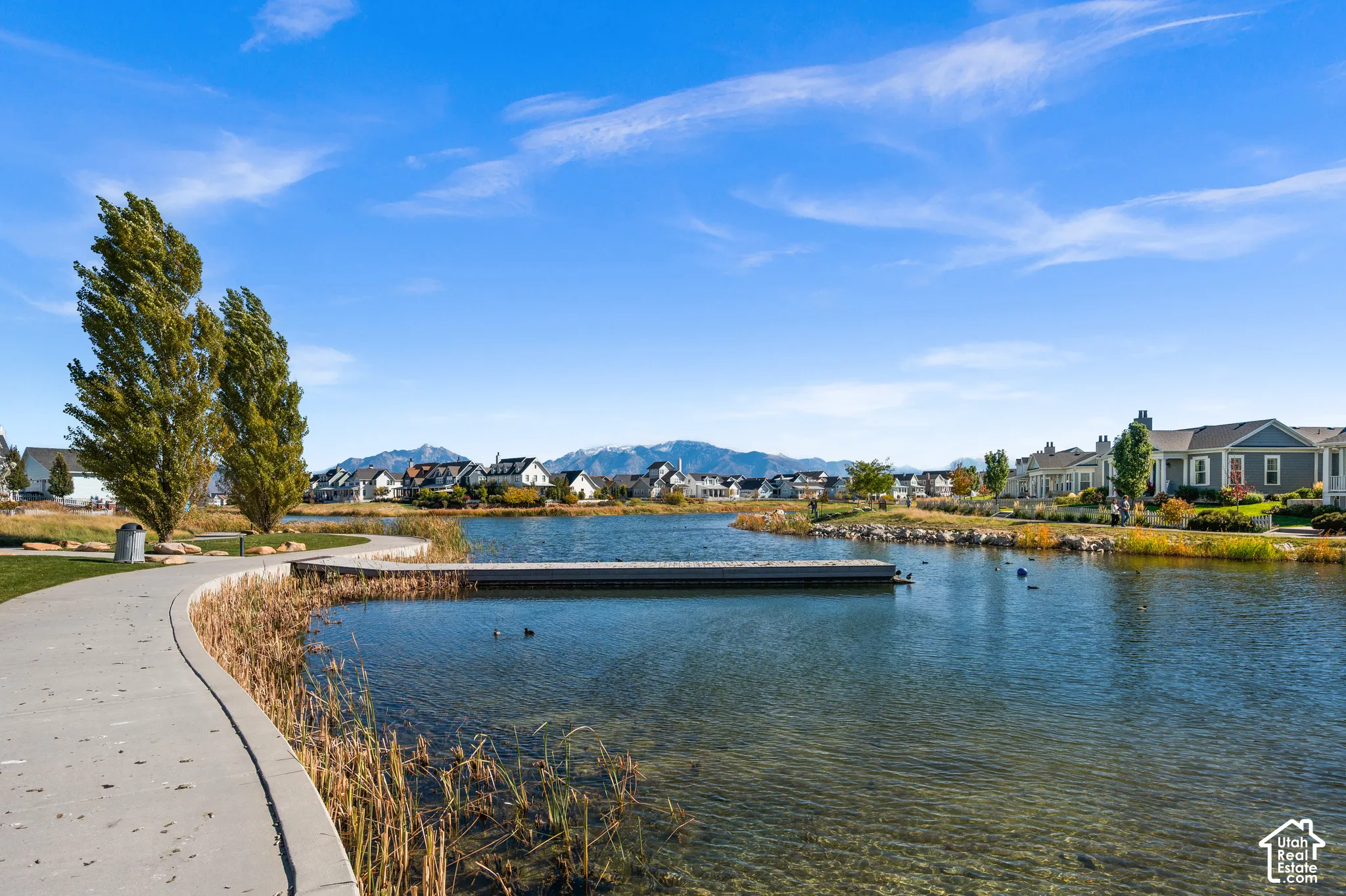 Waterfront community setting featuring a concrete walking path, a wooden pier extending over the pond, native landscaping, and mountain scenery
