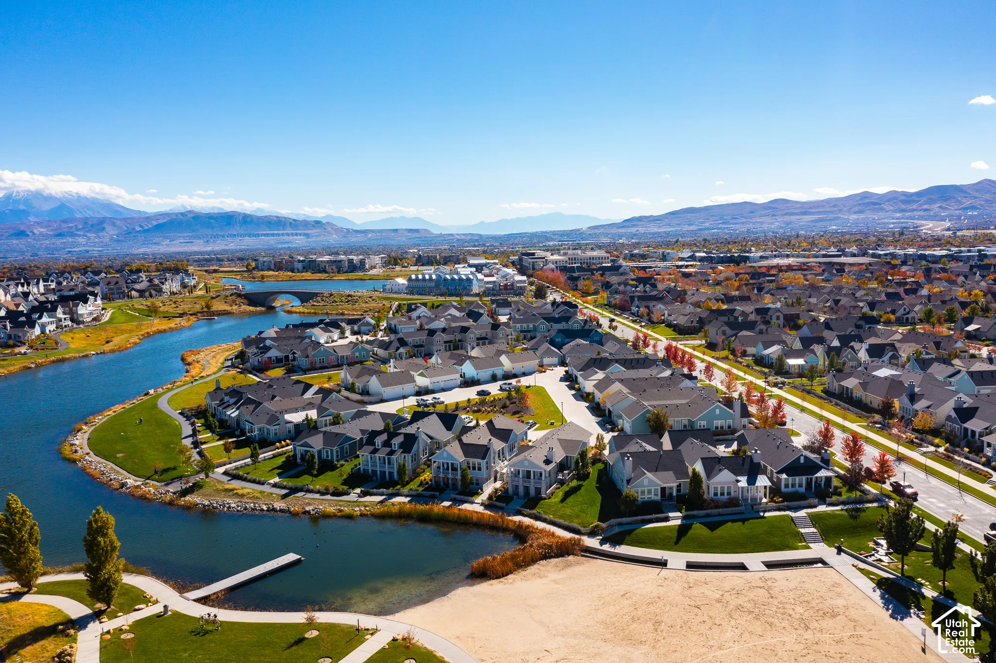 Aerial perspective of a lakeside community featuring direct water frontage, a private dock, sandy beach area, paved walking paths, and a mountainous backdrop
