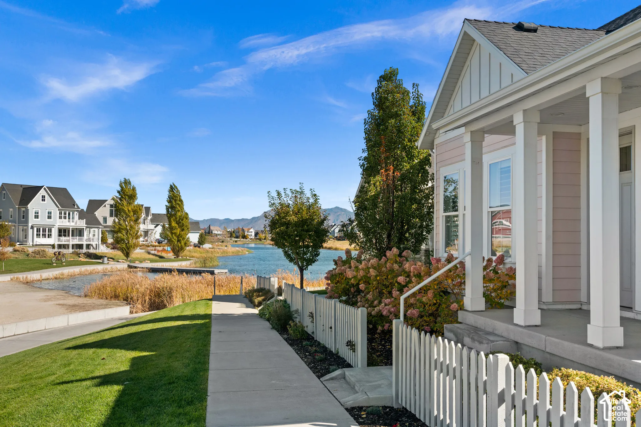Covered front porch featuring white square columns, clapboard siding, gable roofline, and picket fencing, situated along a paved walkway with water frontage and a mountain backdrop