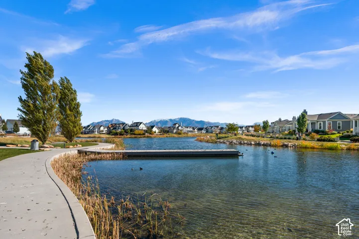 Waterfront community setting featuring a concrete walking path, a wooden pier extending over the pond, native landscaping, and mountain scenery