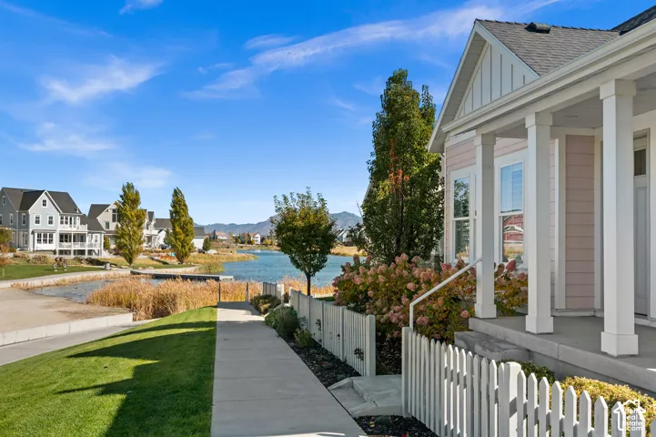 Covered front porch featuring white square columns, clapboard siding, gable roofline, and picket fencing, situated along a paved walkway with water frontage and a mountain backdrop