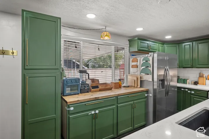 Kitchen featuring green shaker-style cabinetry, wood-finish countertops, stainless steel refrigerator, recessed lighting, and white stone-finish island countertop with integrated sink