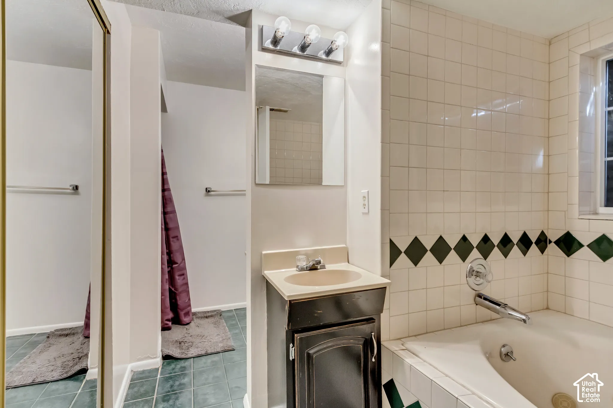 Bathroom featuring a vanity with integrated sink, three-light wall-mounted fixture, recessed wall mirror, tiled soaking tub surround with decorative accent border, and tile flooring