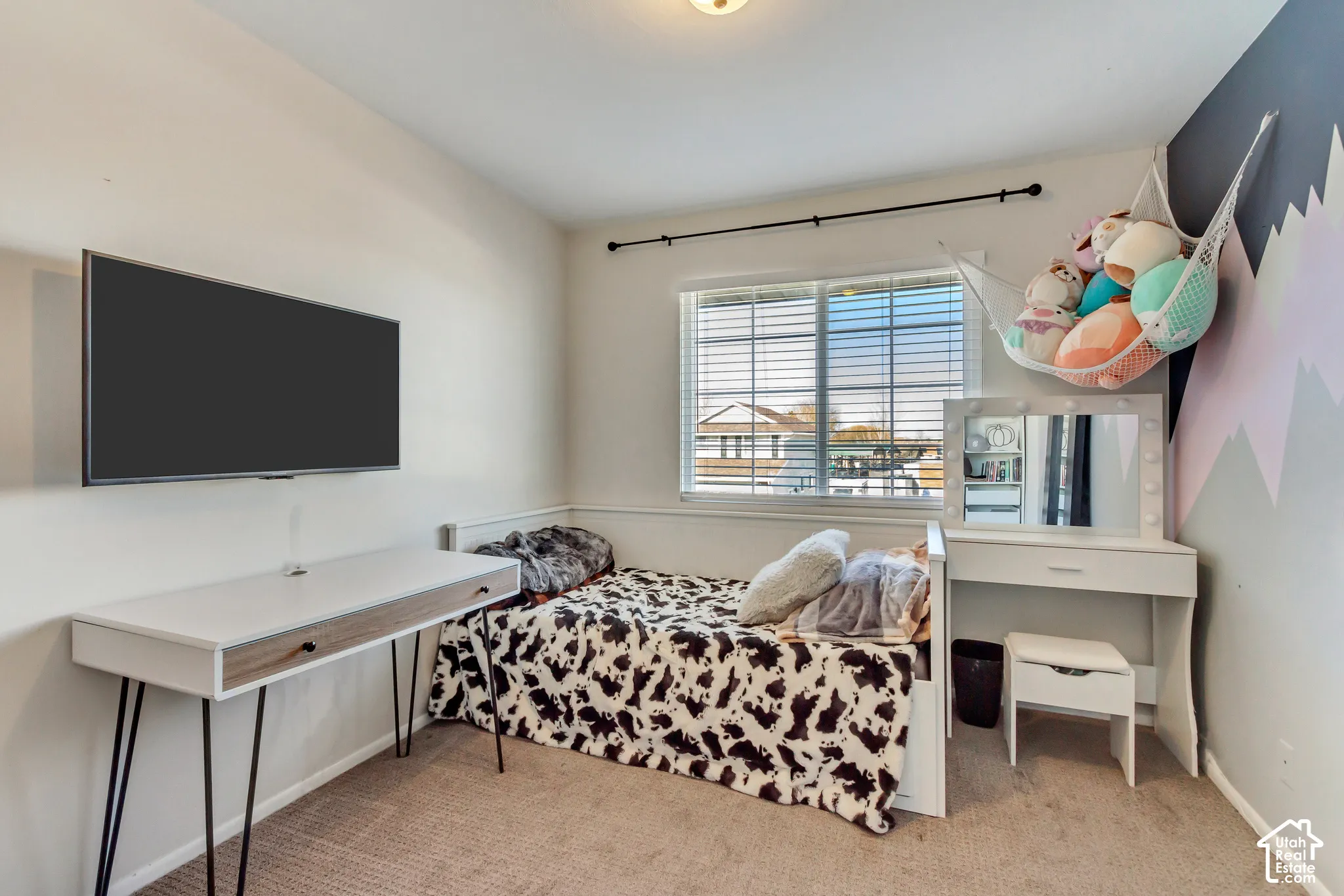 Room featuring neutral-toned carpeting, a wall-mounted flat-screen television, a single horizontal window with integrated blinds, and an accent wall with mountain-inspired wall art