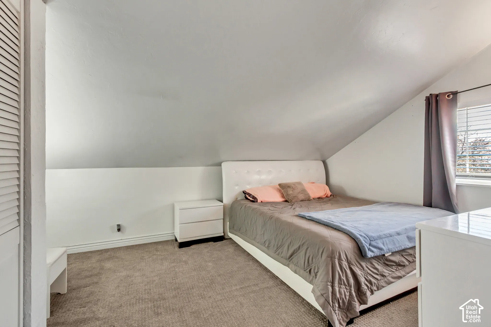 Vaulted ceiling room featuring neutral-toned carpeting, louvered closet doors, and a window with horizontal blinds