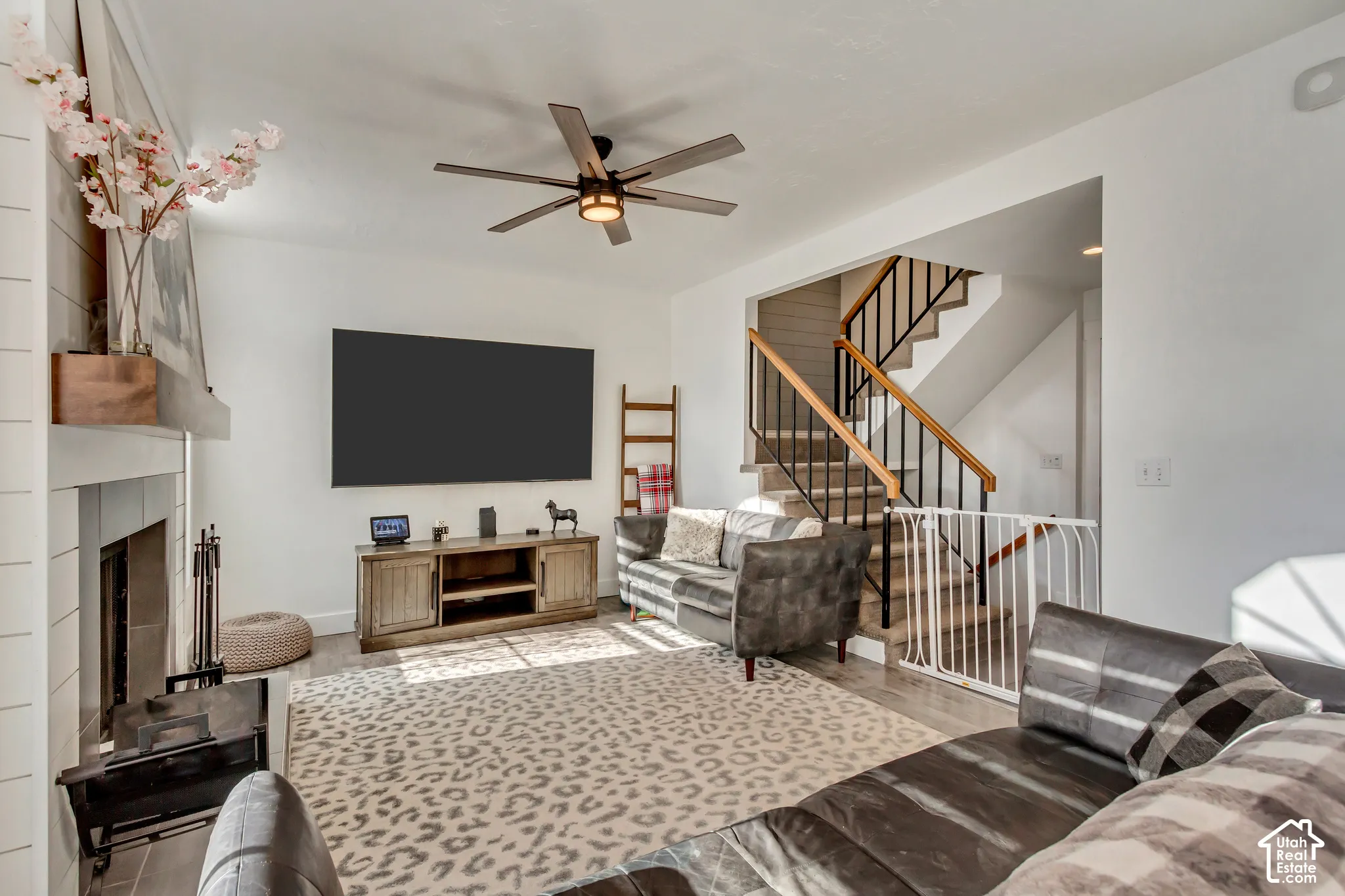 Living area featuring a fireplace with a horizontal shiplap surround, wood-finish flooring, a multi-blade ceiling fan, and a staircase with a wooden handrail and black metal balusters