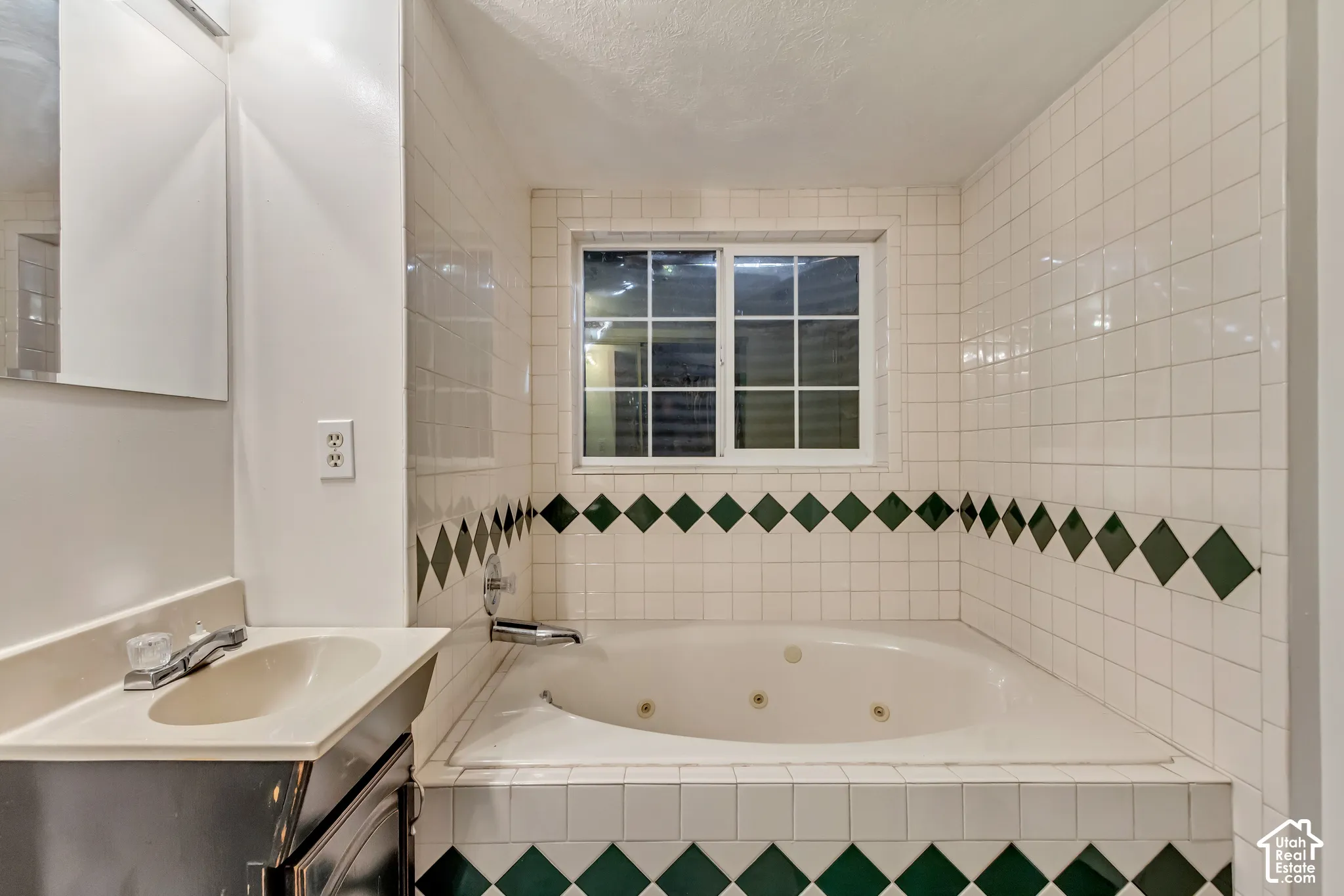 Built-in jetted soaking tub featuring white tile surround with green diamond accent inlay, integrated basin vanity, and grid-pattern window
