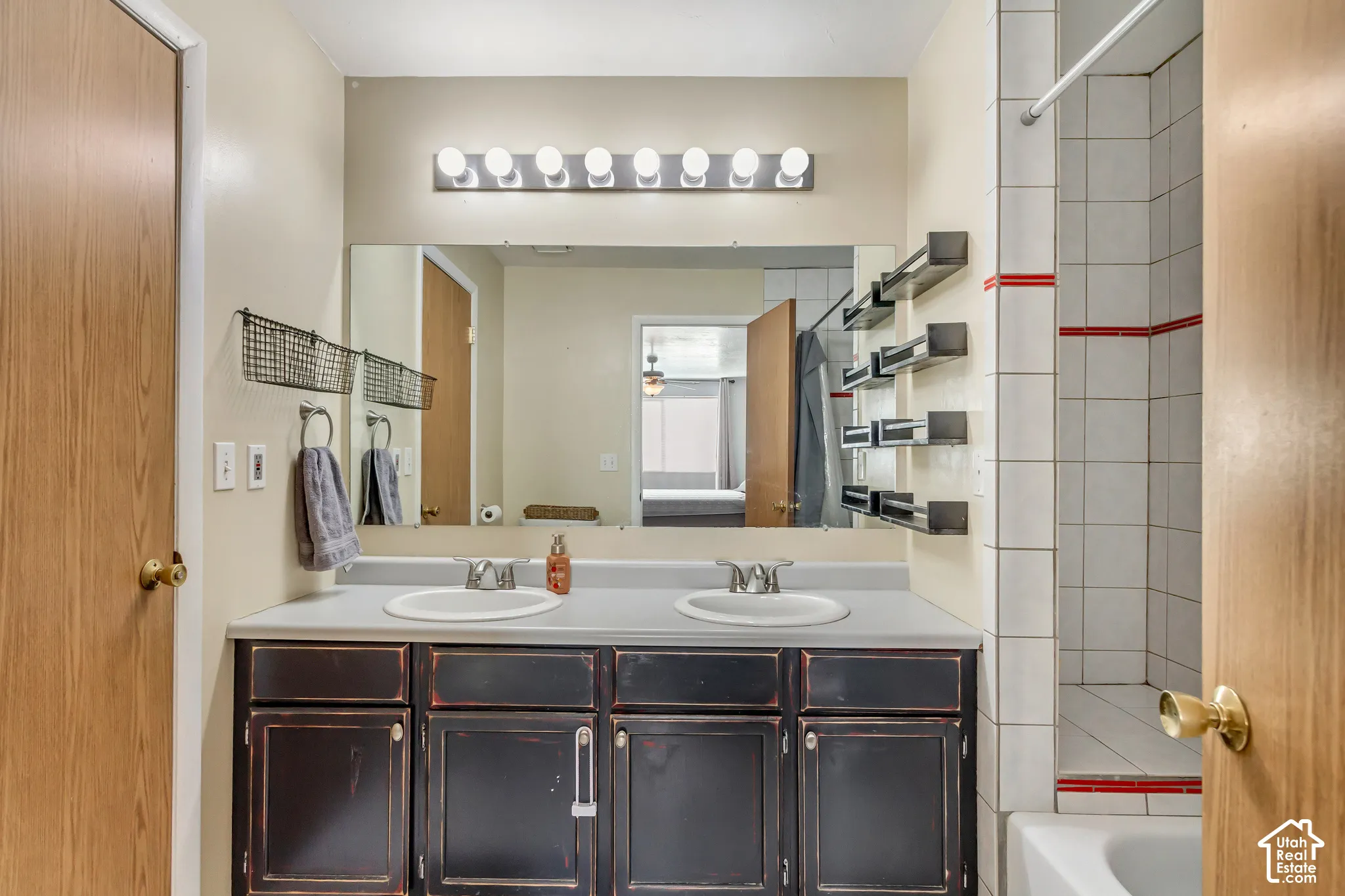 Dual-basin vanity with a solid-surface countertop, full-width wall mirror, eight-bulb horizontal light fixture, and tiled tub-shower combination