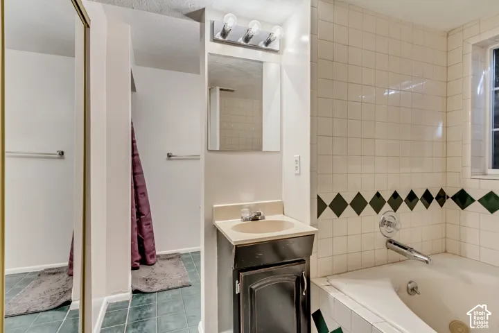 Bathroom featuring a vanity with integrated sink, three-light wall-mounted fixture, recessed wall mirror, tiled soaking tub surround with decorative accent border, and tile flooring