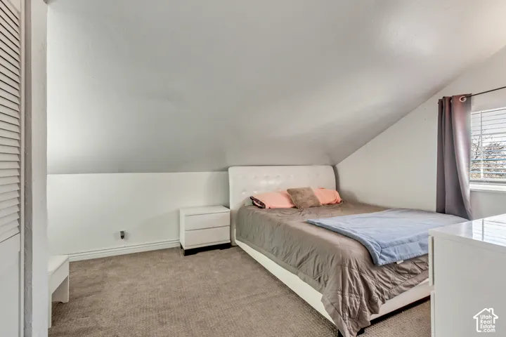 Vaulted ceiling room featuring neutral-toned carpeting, louvered closet doors, and a window with horizontal blinds