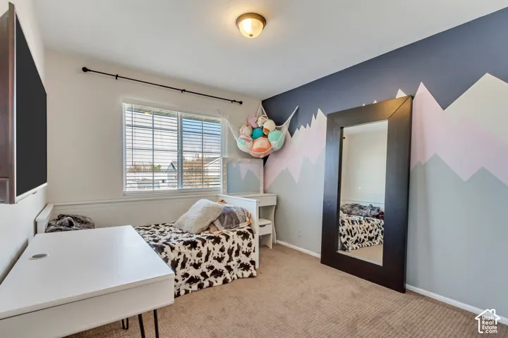 Bedroom featuring neutral carpeting, a dual-pane window with horizontal blinds, a decorative wall mural, and a central flush-mount ceiling light fixture