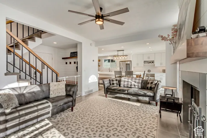 Open-concept interior featuring a fireplace with a wood mantel, staircase with metal railing, ceiling fan with integrated lighting, recessed lighting, and kitchen cabinetry with a central island