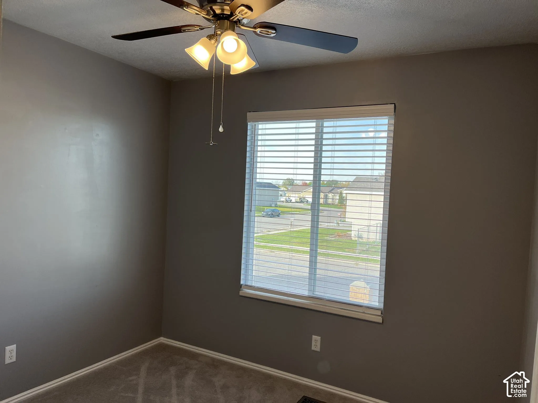 Neutral grey interior walls, ceiling fan with three-light fixture, single-panel window with integrated blinds, and carpeted flooring