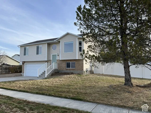 Split-level exterior featuring light-toned horizontal siding, brick masonry, an attached two-car garage, white vinyl fencing, and a mature evergreen tree