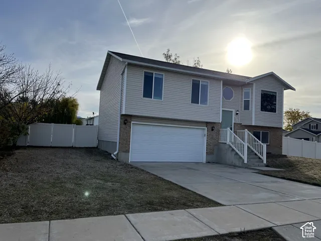 Split-level exterior featuring light-toned horizontal siding, brick masonry accent, two-car garage, concrete driveway, and white vinyl privacy fencing