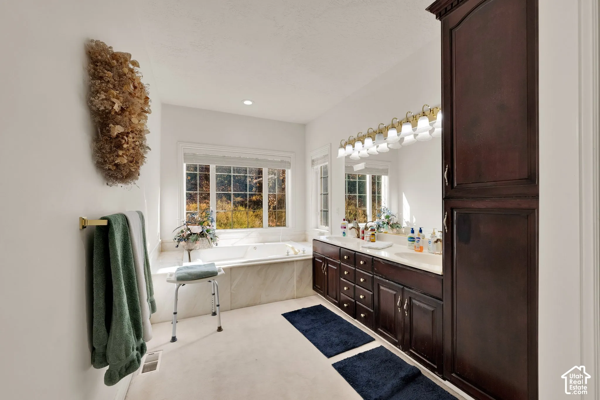 Bathroom featuring dark wood-finish cabinetry, built-in tall storage, dual-sink vanity with a countertop, oversized soaking tub with tile surround, and gold-finish hardware
