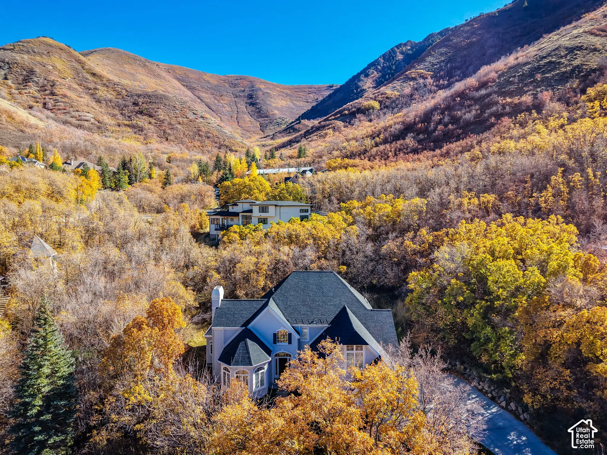 Multi-level residence featuring a dark shingle roof, turret-style architecture, light-toned siding, and exterior chimney, set against a mountainous backdrop with mature deciduous trees