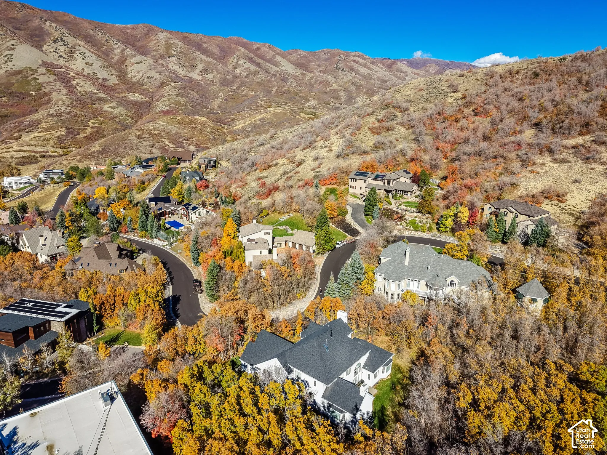 Aerial perspective of a hillside residential community featuring multi-level dwellings, asphalt roads, established trees with autumn foliage, and a mountain range backdrop