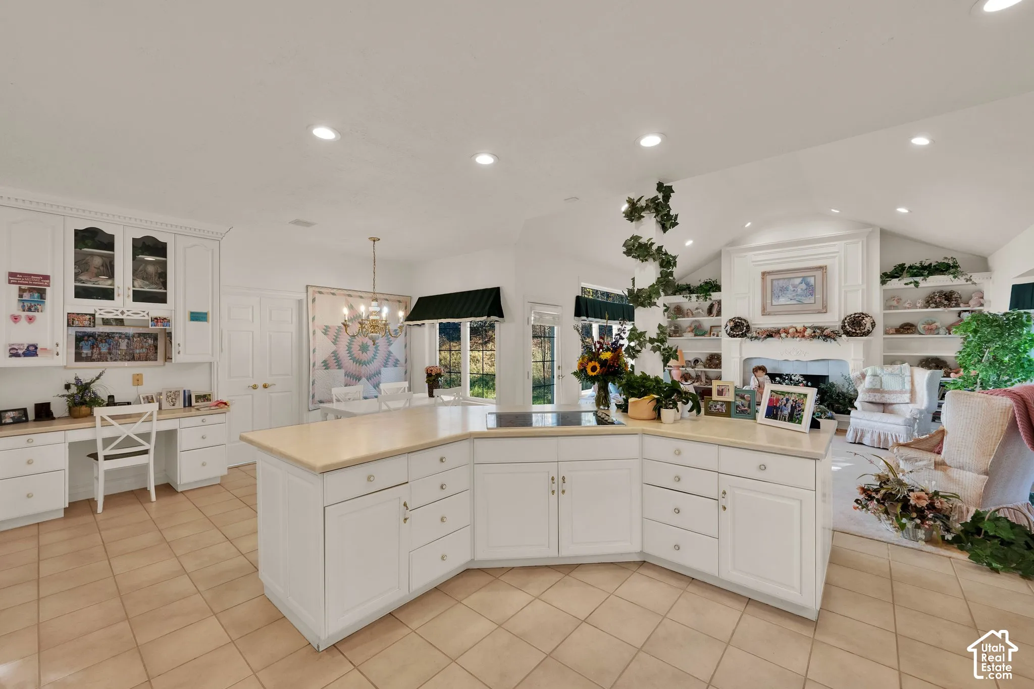 Kitchen island with integrated cooktop and neutral stone-finish countertops, custom white cabinetry with built-in desk area, recessed lighting, and a fireplace featuring a detailed mantel and built-in shelving